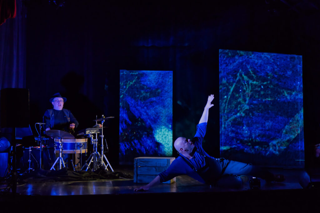 Scène du spectacle avec un batteur dans le fond et un comédien en premier plan dans un décor entre bleu et noir.